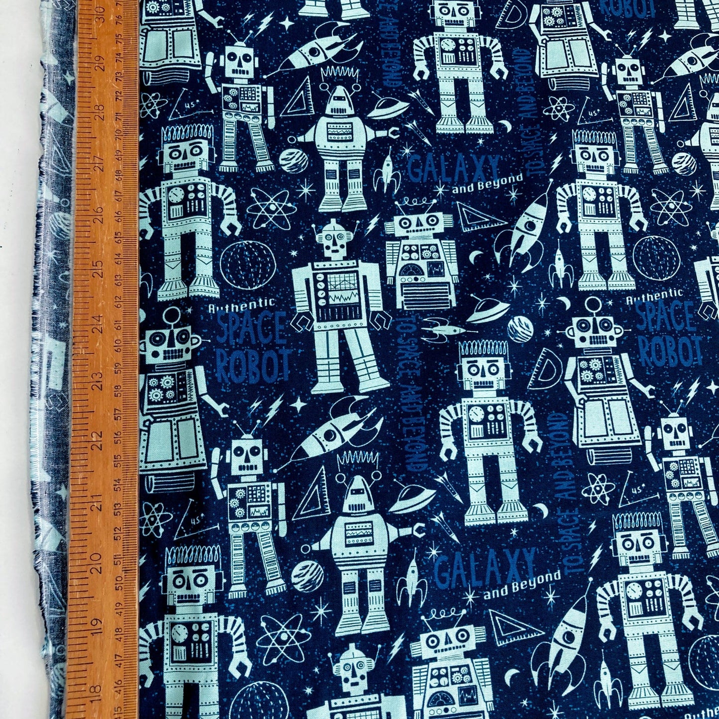 Quilting cotton fabric with robot and space-themed pattern on a blue background, measured with a ruler.