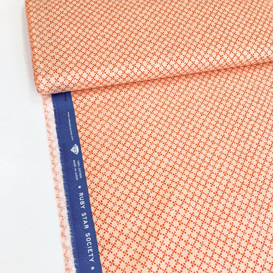 A bolt of quilting cotton fabric with an all-over tile print in terracotta on a peach background, with contrasting blue Ruby Star Society selvedge