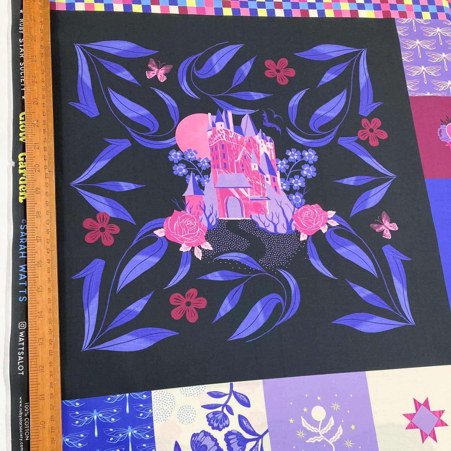 Quilting cotton panel showing the largest illustraions: a fairy castle on a black background, surrounded by purple leaves and pink flowers, with a ruler for scale.