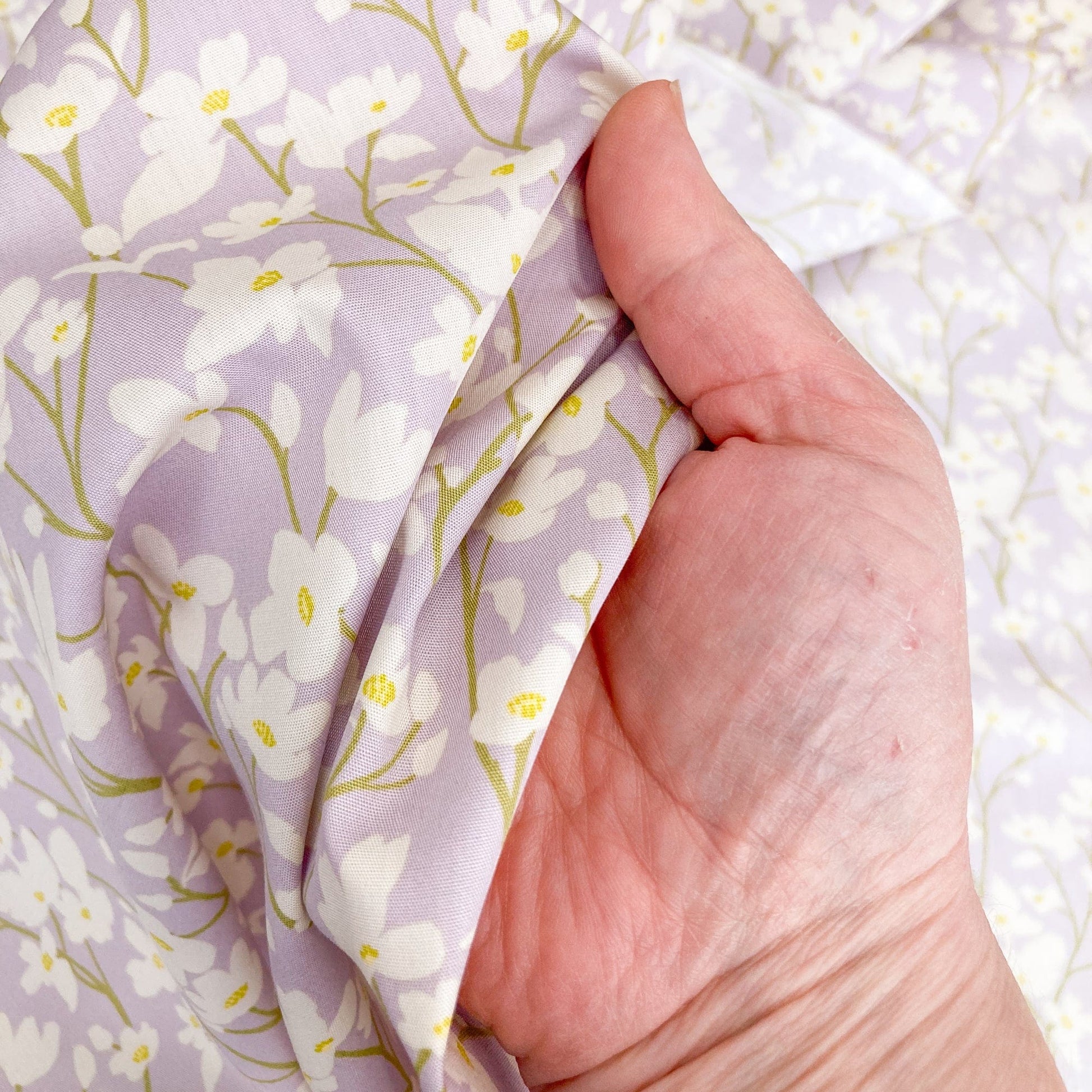 Hand holding a floral cotton fabric with cream flowers on a lilac background.