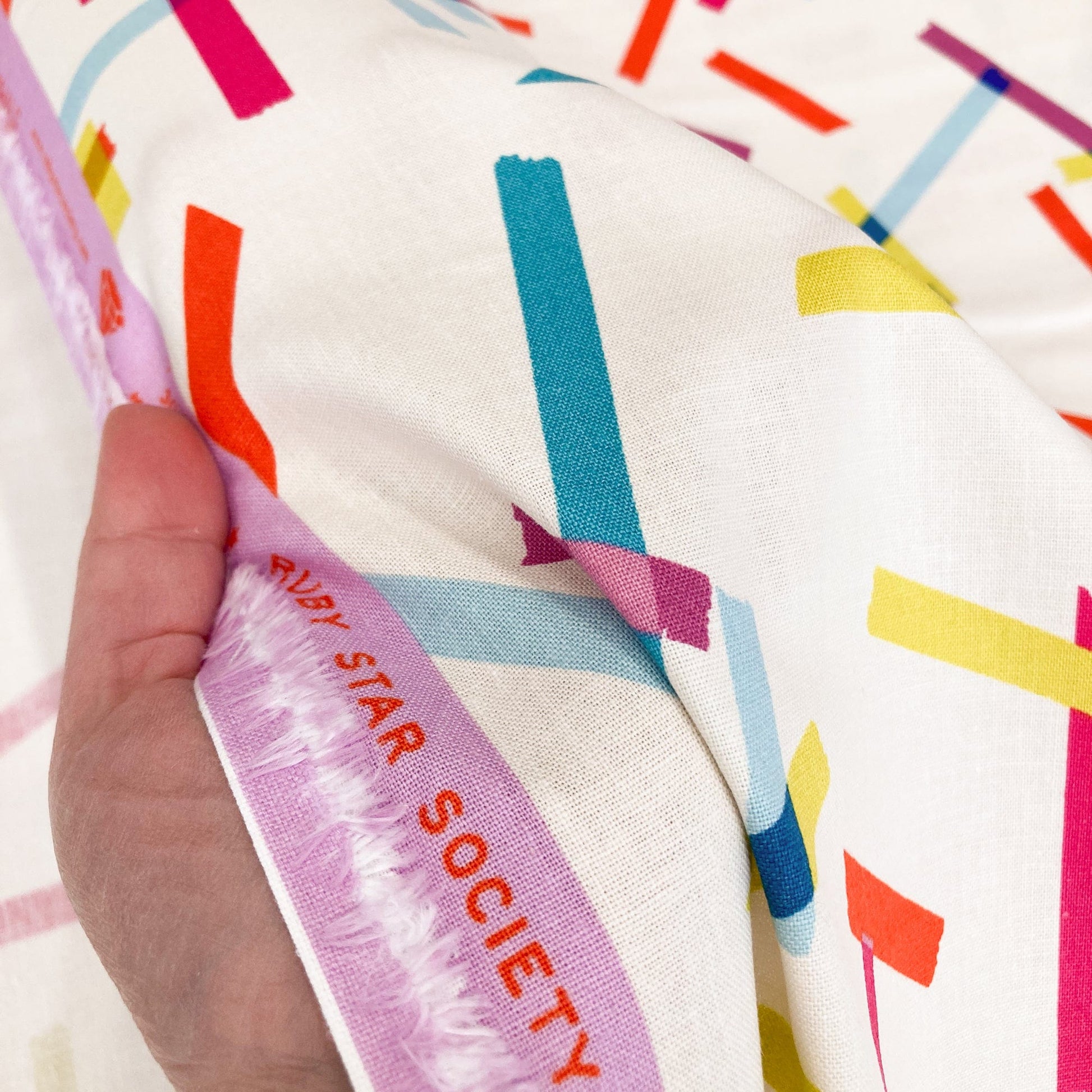 Cream cotton & linen canvas fabric with washi tape print in bright colours held by a hand.