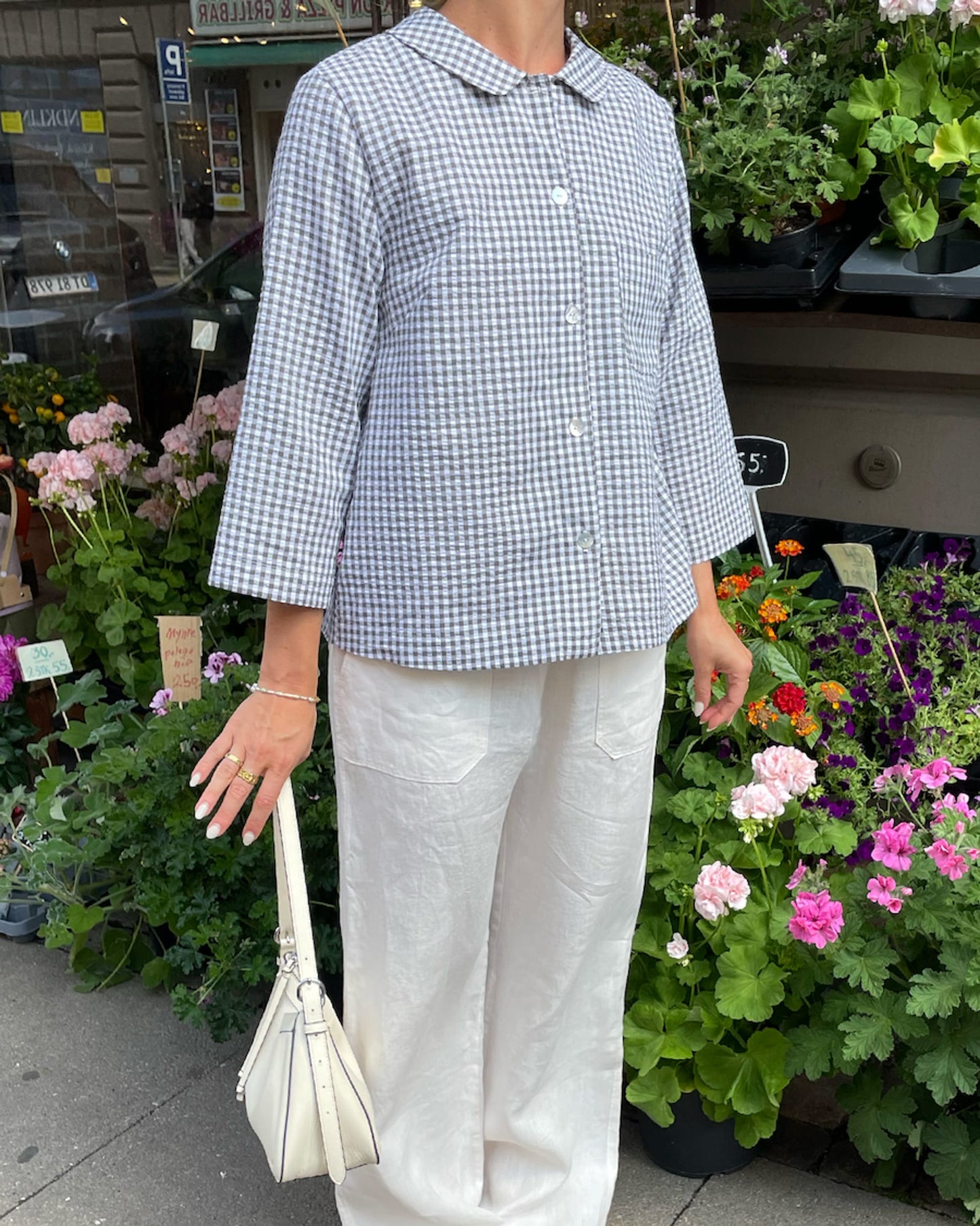Person wearing a blue and white checkered shirt and white pants, standing among flowers and plants.