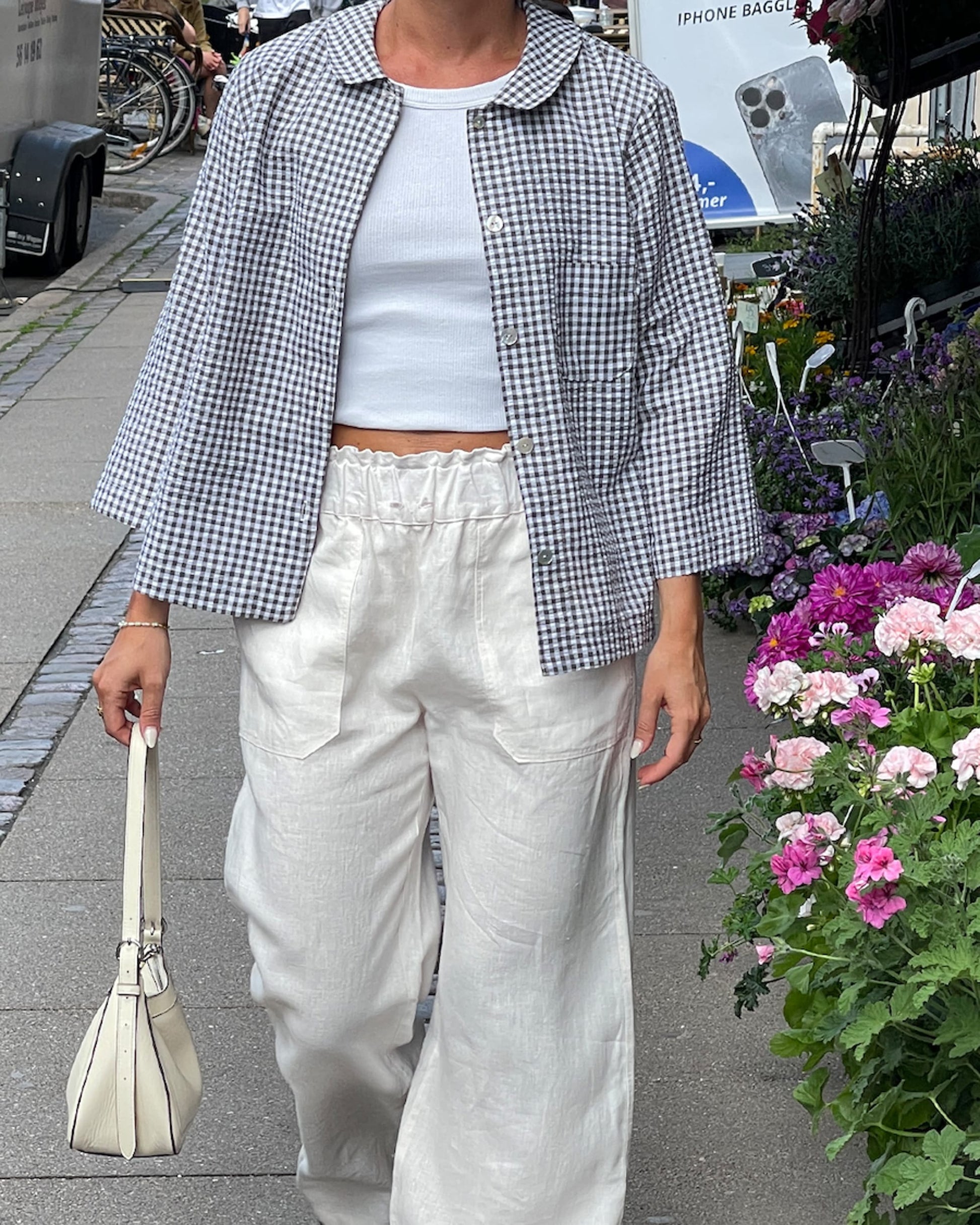 Person wearing a checkered shirt, white pants, and holding a beige bag on a street with flowers in the background.